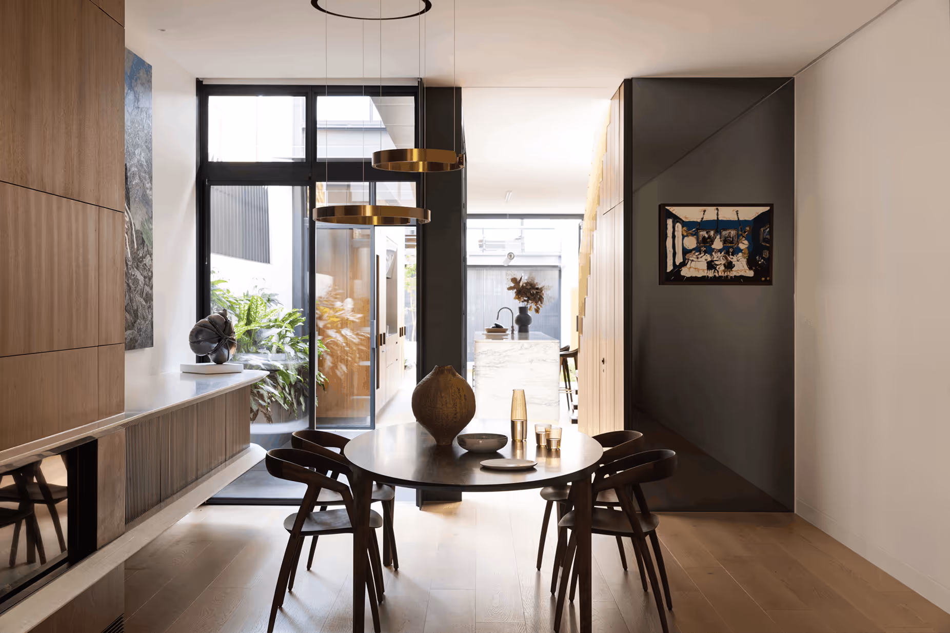 Modern dining area with a round table, four wooden chairs, minimalist decor, and large windows overlooking greenery.