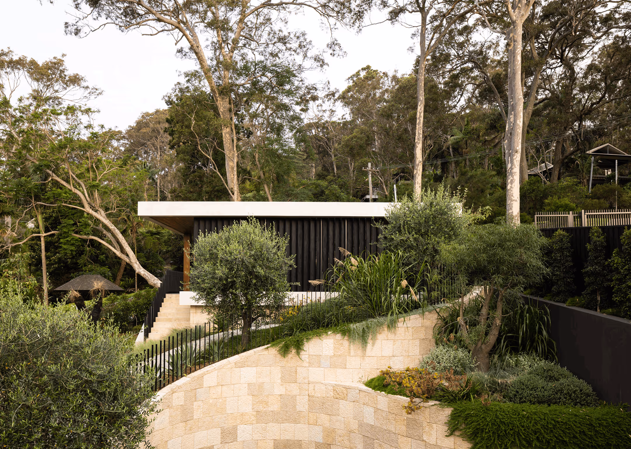 Modern house with a flat white roof surrounded by lush greenery and tall trees on a sloped stone retaining wall.
