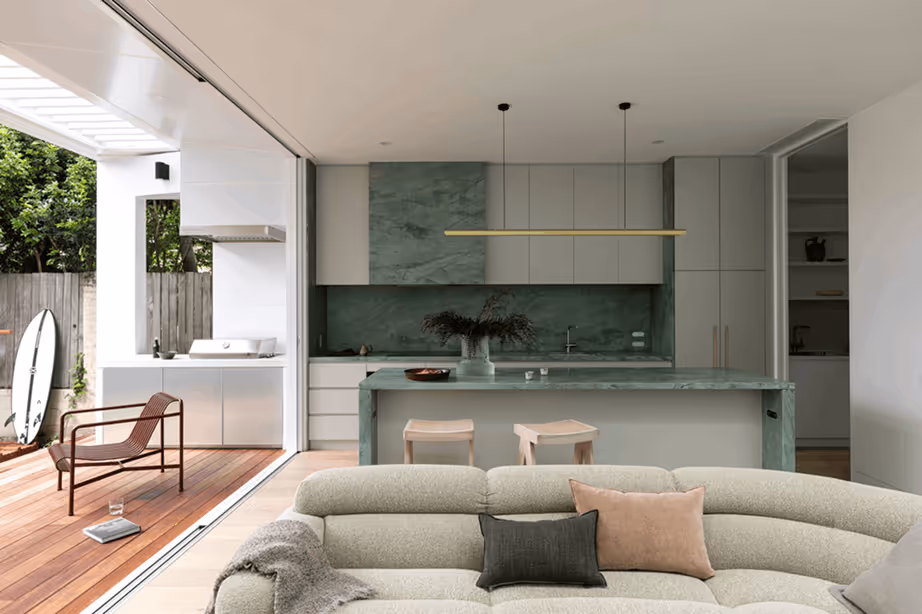 Modern open-concept living space with a curved beige sofa in the foreground, a green marble kitchen island with two stools, and sliding doors opening to an outdoor patio with a chair and surfboard.