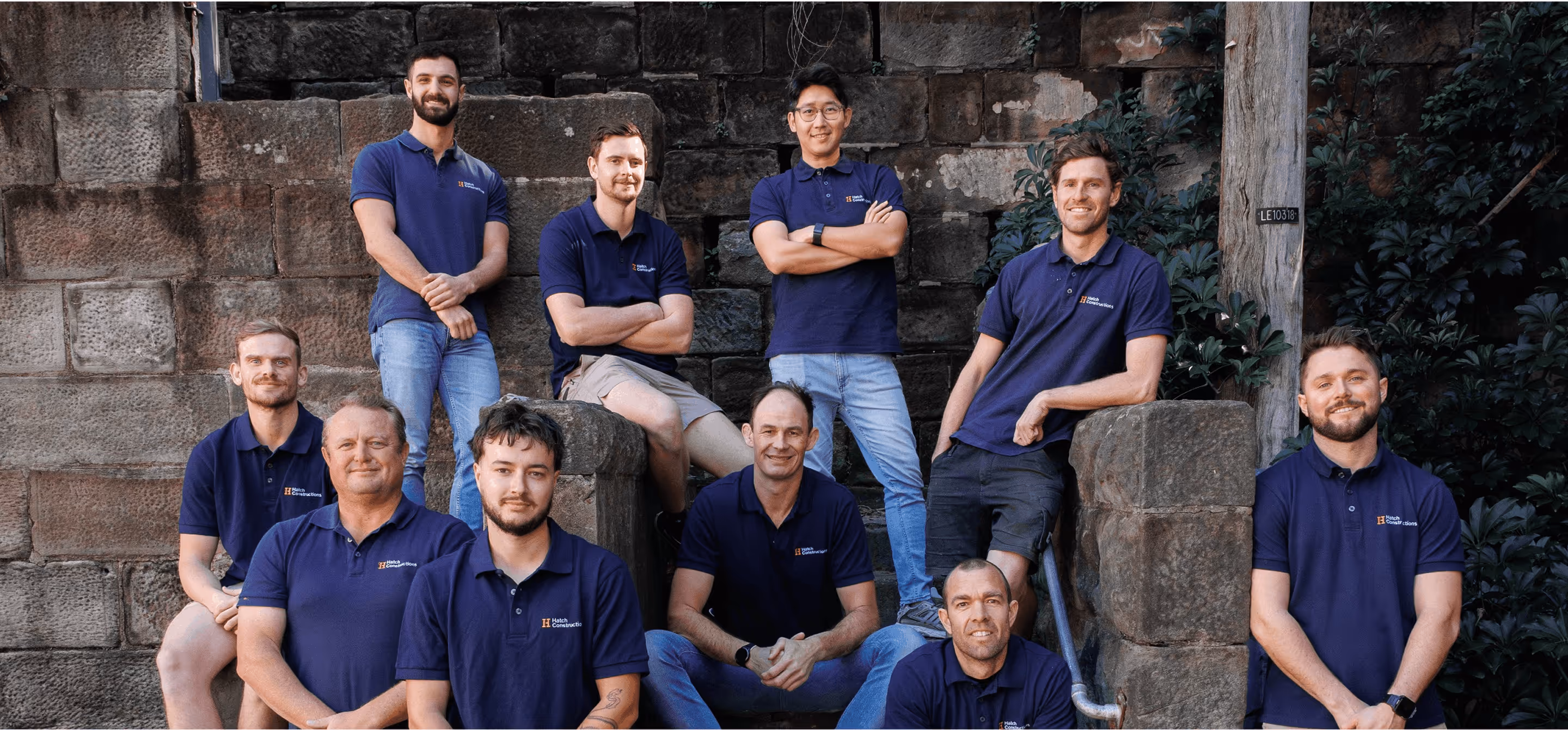 Group of ten men wearing matching navy blue polo shirts posing outdoors against stone walls and greenery.