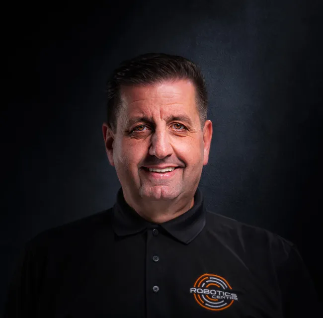 Middle-aged man smiling in a black Robotics Center polo shirt against a dark background.