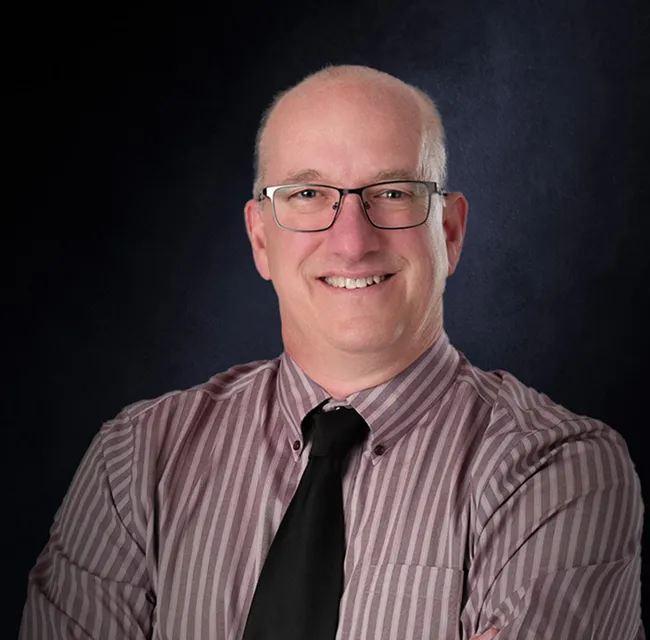 Smiling bald man with glasses wearing a striped shirt and black tie against a dark background.