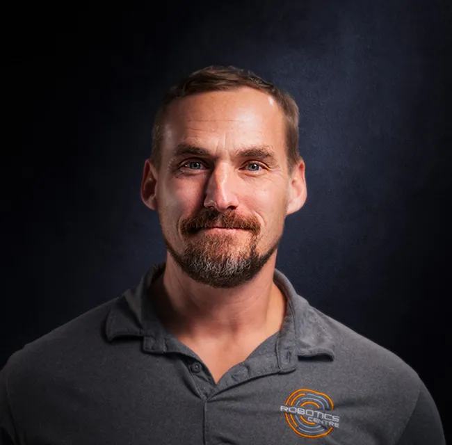 Portrait of a man with short hair and a beard wearing a grey polo shirt with a Robotics Centre logo on a dark background.