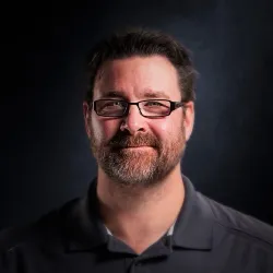 Smiling man with glasses, beard, and short hair wearing a dark shirt against a dark background.