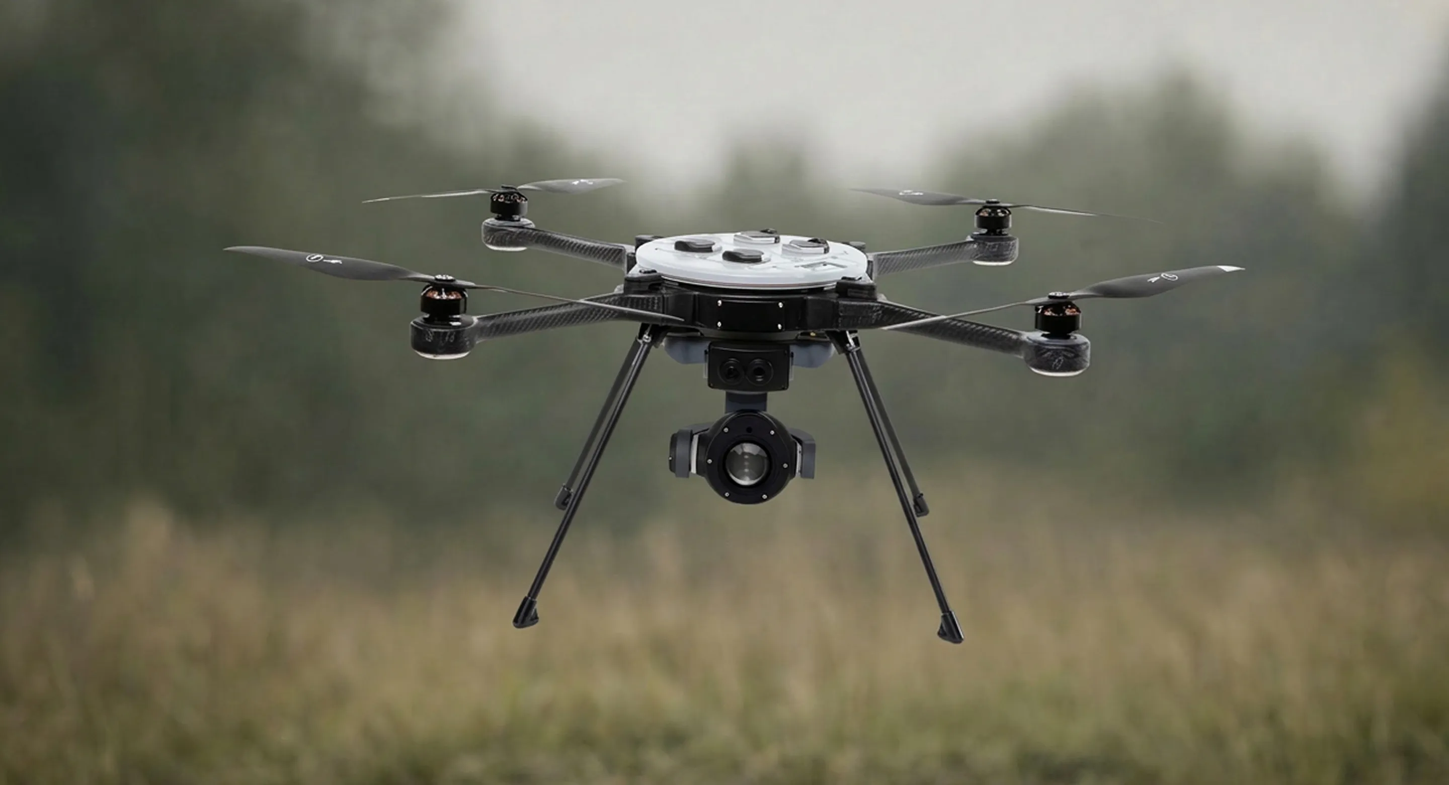 Black drone with six propellers and a camera hovering over a grassy field with blurred trees in the background.