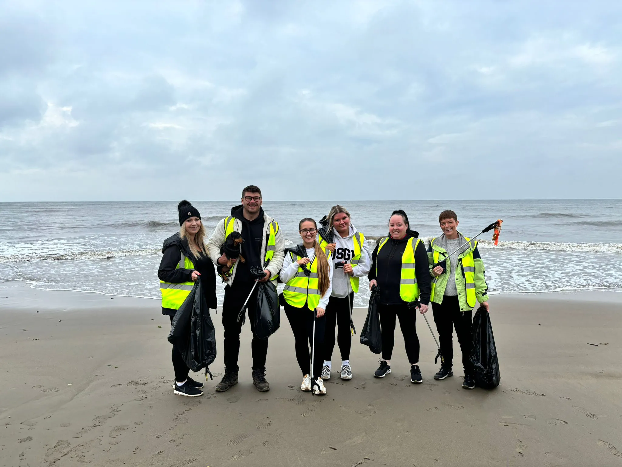 Group of six people wearing yellow safety vests standing on a beach with trash bags and litter pickers during a cleanup.