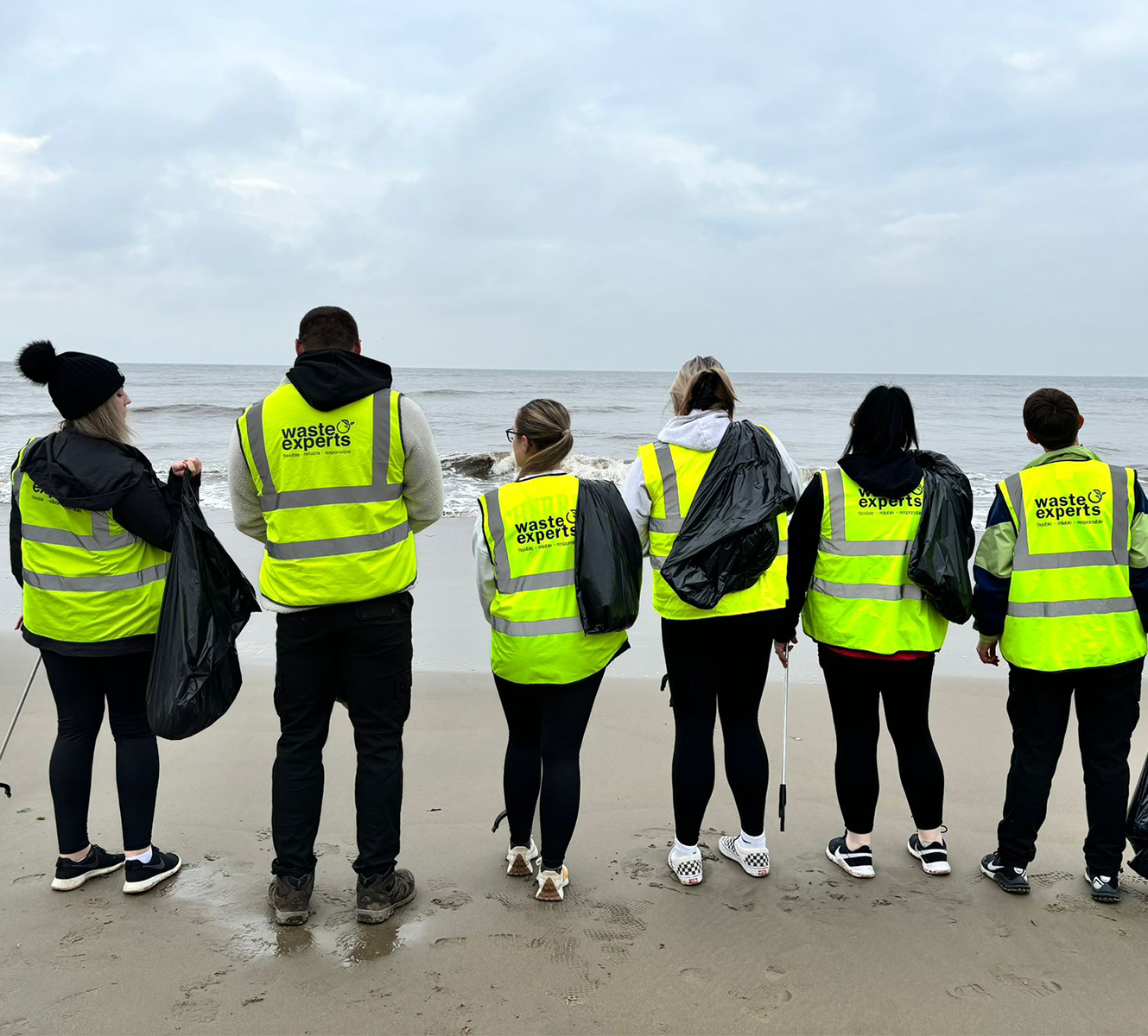 Six people wearing yellow 'waste experts' vests standing in a line on a beach holding black trash bags facing the ocean on a cloudy day.