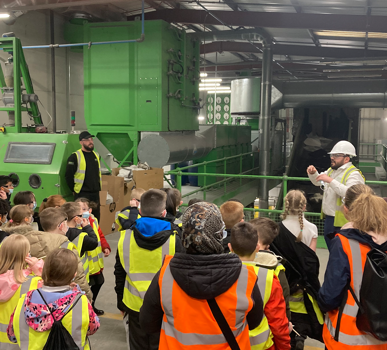 Group of children wearing high-visibility vests on a factory tour with two adults, one explaining near green industrial machinery.