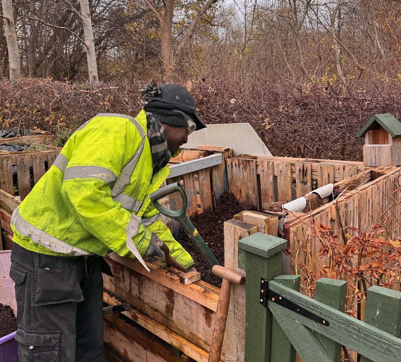 Person in a bright yellow jacket working with compost in wooden bins outdoors during late autumn.