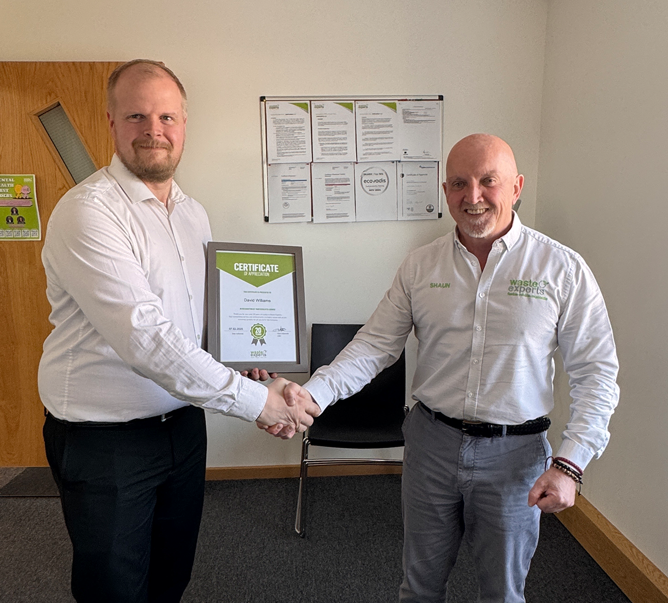 Two men shaking hands, one holding a framed certificate of appreciation, standing in an office with documents on the wall behind them.