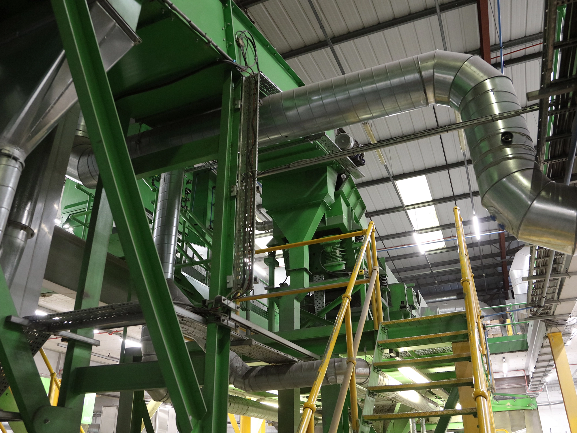 Industrial facility interior with green metal framework, yellow safety railings, and large silver ventilation ducts.