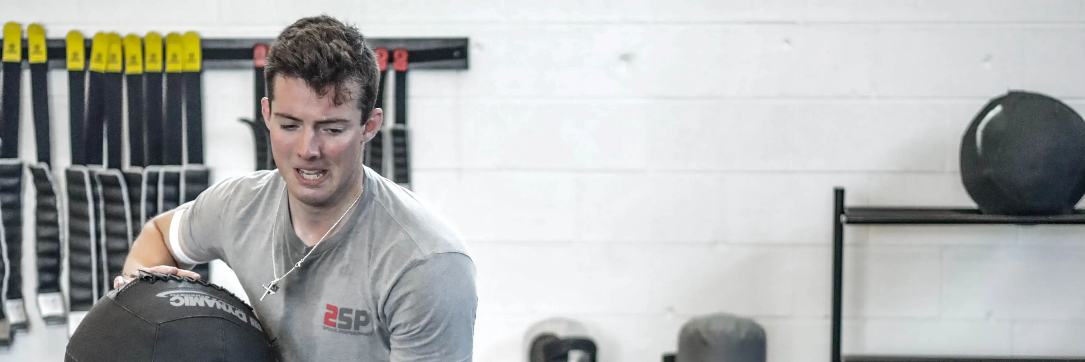 Man in gray 2SP Sports Performance shirt lifting a black dynamo medicine ball in a gym.