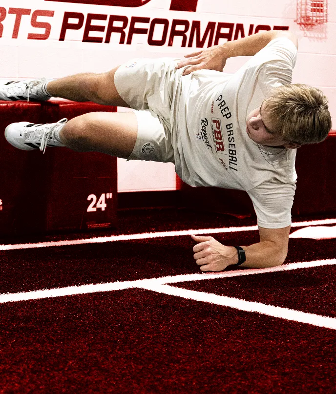 Male athlete wearing white sportswear performing a side plank exercise on a maroon indoor turf with white lines.
