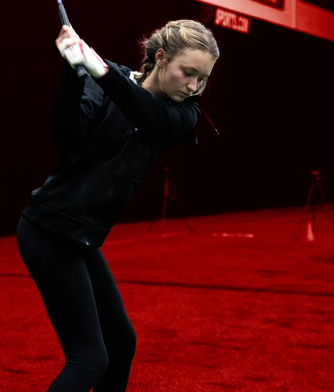 Woman in black athletic clothing practicing a golf swing indoors on red turf.