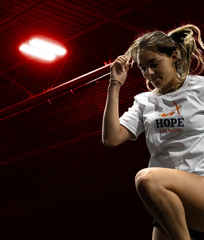Young woman in white Hope Lacrosse shirt and white sneakers performing a high knee exercise on a red floor with a dark background.