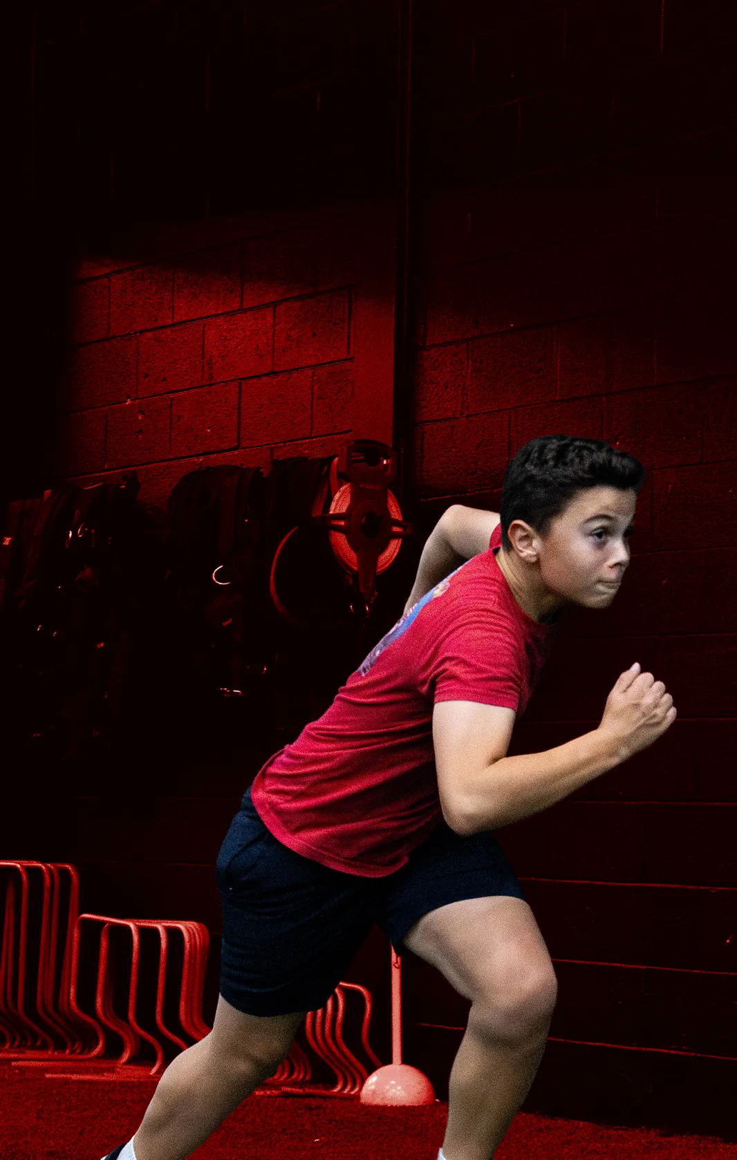 Young boy in red shirt and black shorts sprinting indoors on a red turf floor.