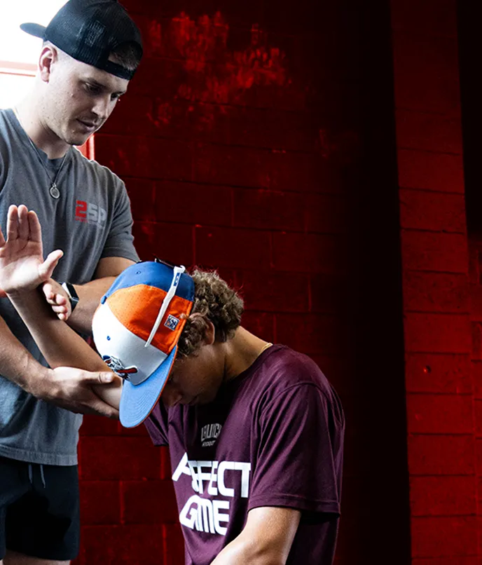 A man in a gray shirt and black cap assists a young athlete wearing a colorful cap and maroon shirt with arm stretching in an indoor gym with red walls.