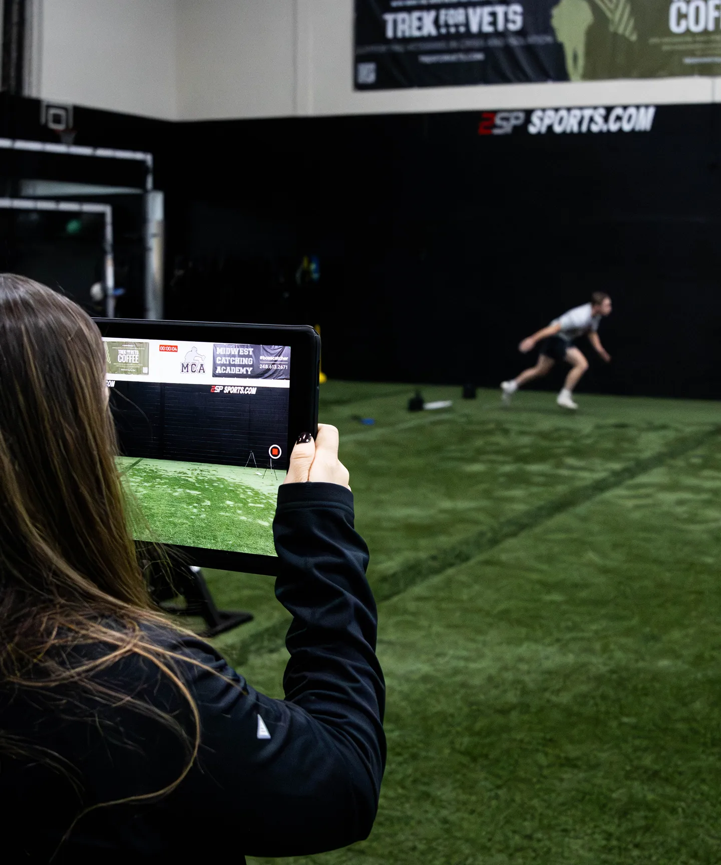 Person recording an athlete sprinting on indoor turf field with a tablet.