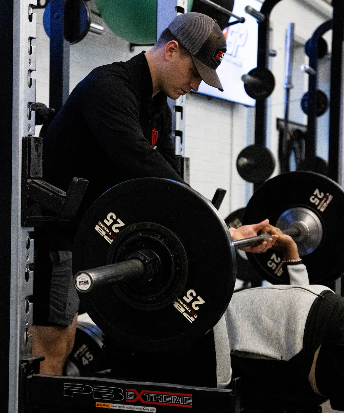 Trainer assists person lifting barbell with 25-pound plates on bench press in a gym.