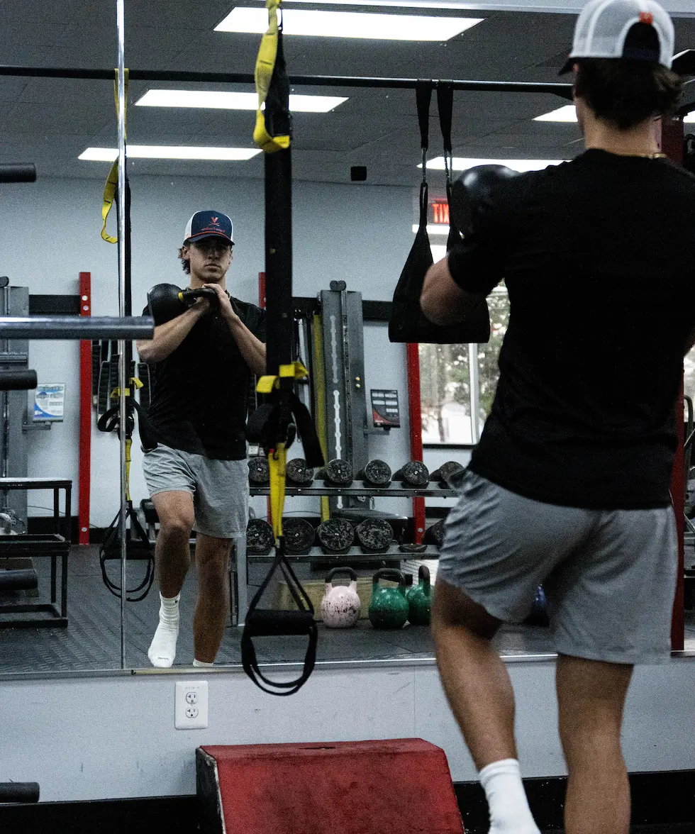 Young man in gym wearing a baseball cap and gray shorts lifts a kettlebell while standing in front of a mirror.