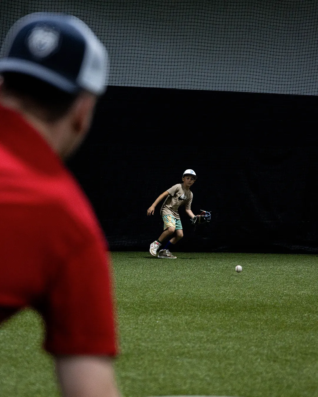 Young boy in baseball gear preparing to catch a rolling baseball indoors with a person in a red shirt and cap watching in the foreground.