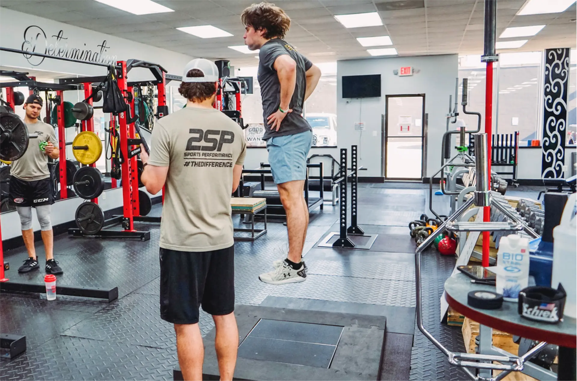 Two men in a gym, one jumping with hands on hips while the other watches holding a stopwatch, surrounded by weightlifting equipment.
