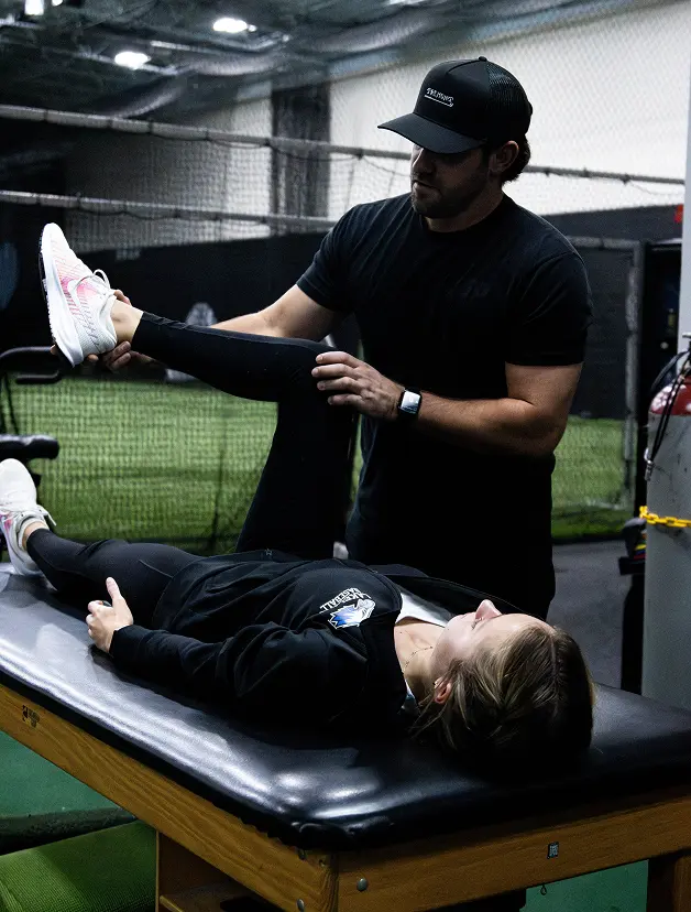 A physical therapist or sports trainer performing assisted hamstring and glute stretching on an athlete lying on a training table.