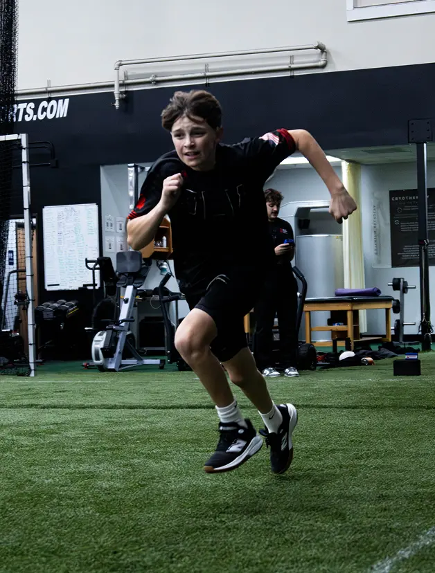 A young athlete sprinting on an indoor turf during a sports performance training session. The individual is wearing a dark top, black shorts, and black-and-white sneakers