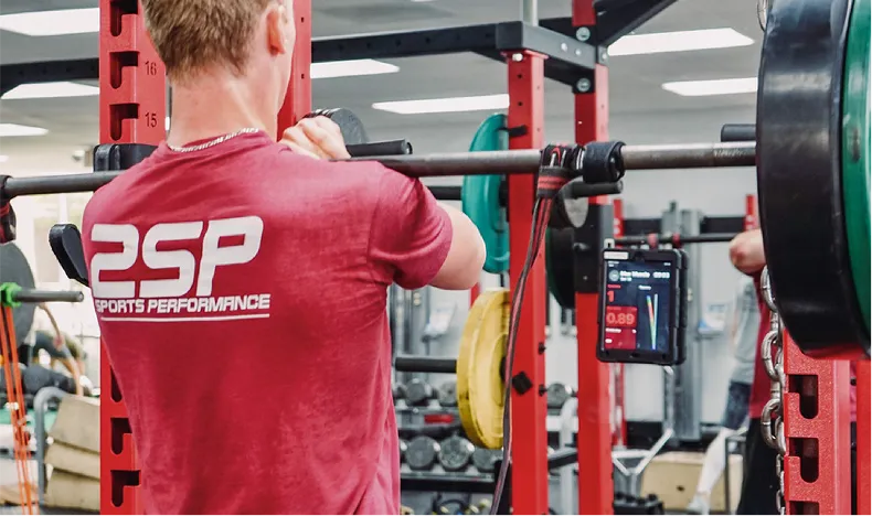 Man in a red DSP Sports Performance shirt lifting a barbell in a gym with weight plates and fitness equipment in the background.