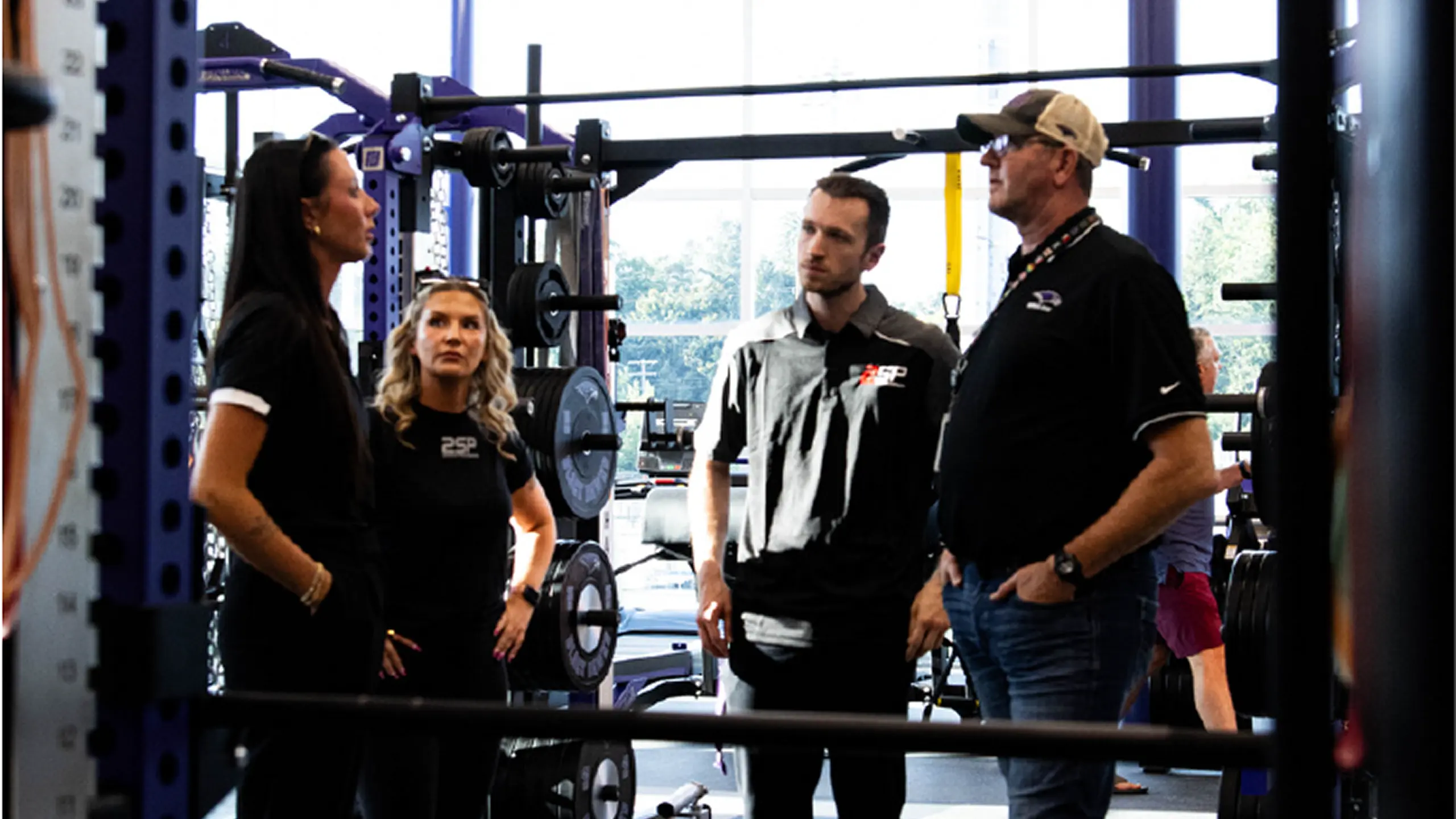 A group of four individuals in a commercial fitness facility, focused around a weight rack and storage system. The two men and two women appear to be engaged in a discussion or training session, surrounded by barbells, weight plates, and functional training equipment.