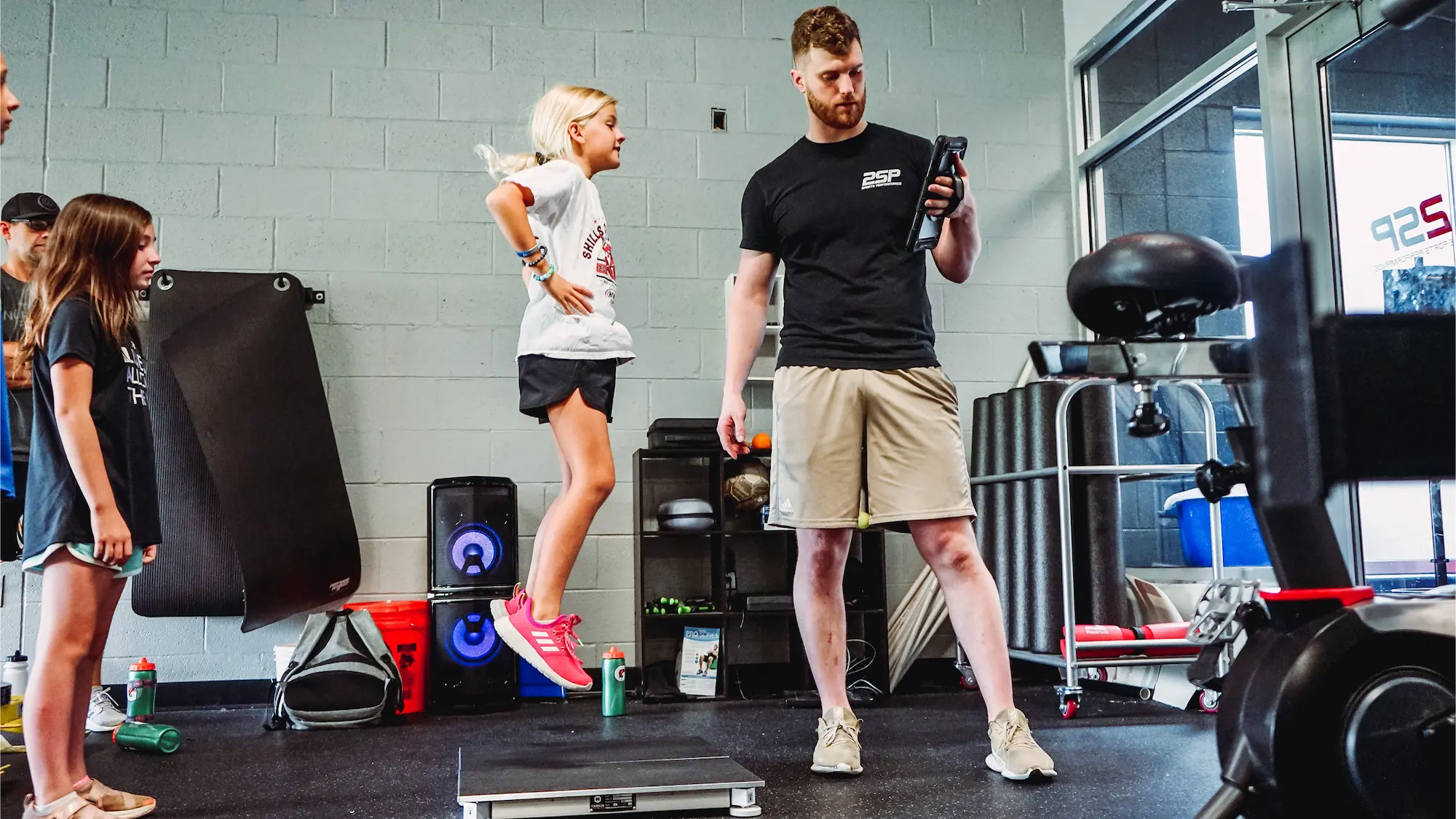 A  youth athletic development session where a coach is guiding a young athlete through a box jump, assessing her performance and power metrics using a digital tablet.