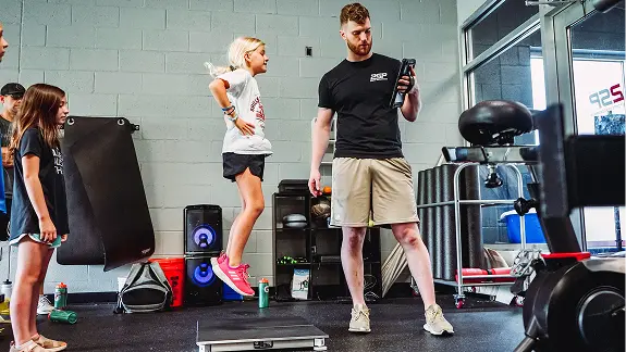 A  youth athletic development session where a coach is guiding a young athlete through a box jump, assessing her performance and power metrics using a digital tablet.
