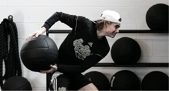 An athlete training at Athlete Lab in Detroit, wearing apparel from the HoneyBaked Hockey Club. They are utilizing black heavy-duty wall balls for a functional strength and conditioning workout.