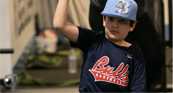 A young baseball player for the Bulls Baseball team, wearing a light blue helmet with a "B" and a navy shirt, caught mid-motion during an athletic activity or training session.