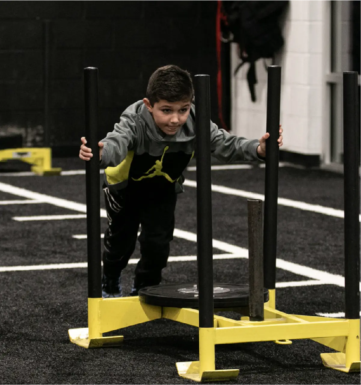 A young boy pushing a weight sled on an indoor turf field, a popular drill in youth sports performance training to build functional leg strength, core stability, and explosiveness