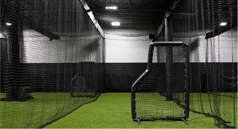 Indoor baseball or softball batting cages with green artificial turf and black protective netting.