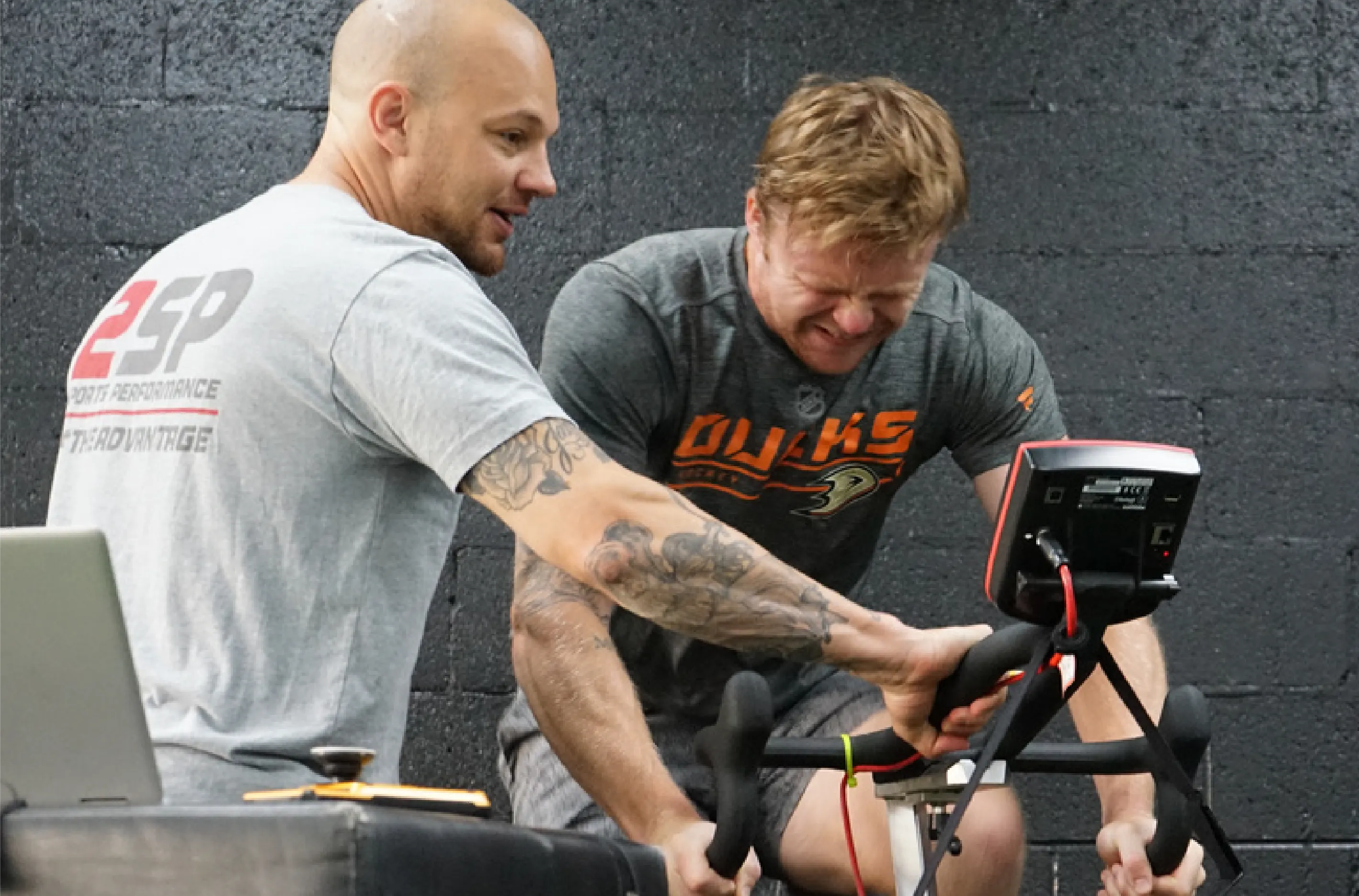 Trainer encourages man struggling intensely on stationary bike during indoor workout session.