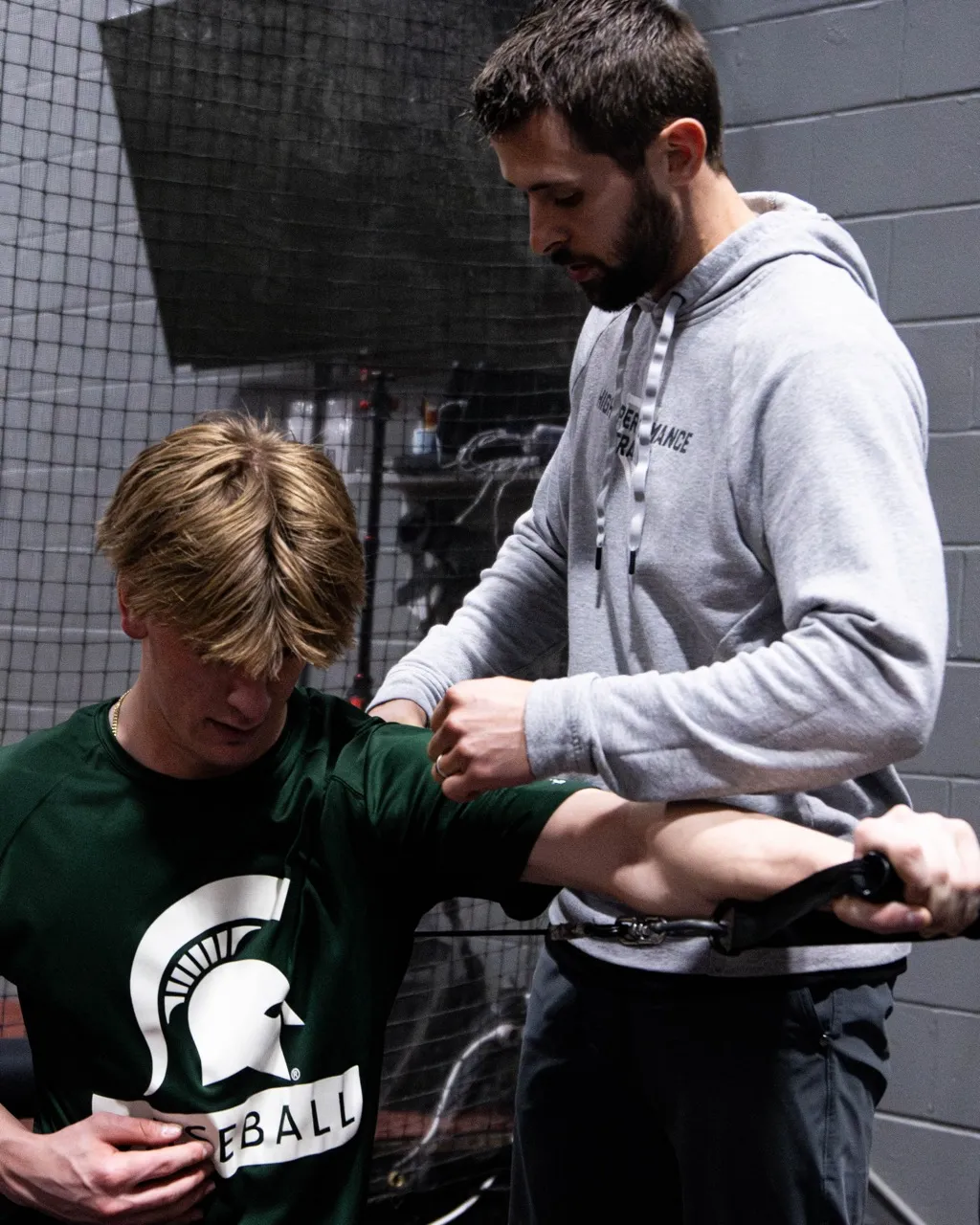 An athlete wearing a Michigan State Spartans logo shirt doing banded shoulder external rotation exercises. This movement isolates and strengthens the rotator cuff muscles.