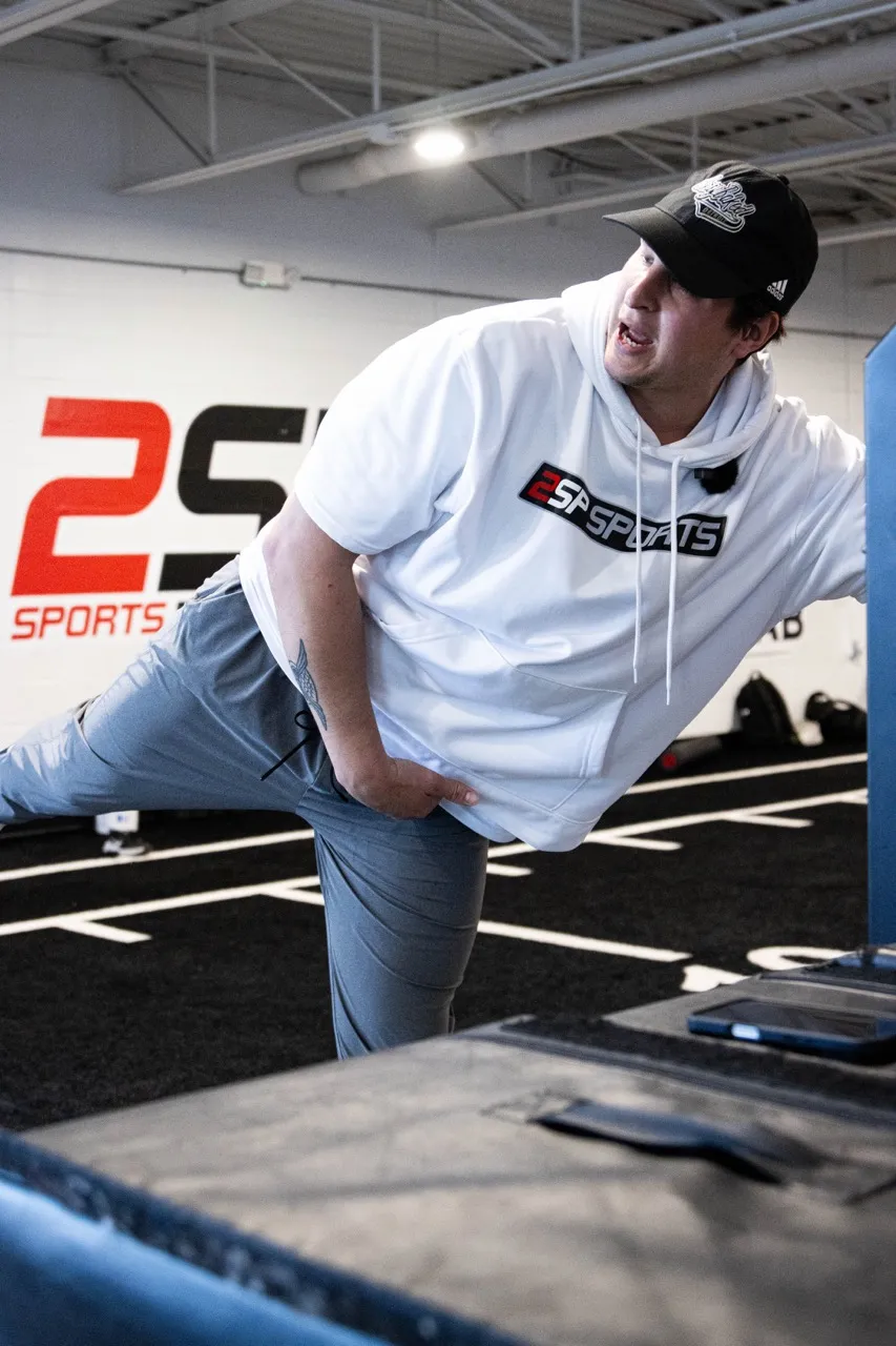 A strength and conditioning coach demonstrating a single-leg hip hinge or rotational movement at 2SPORTS, an athletic performance facility