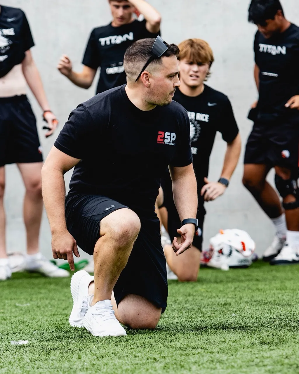 Coach in black shirt kneeling on turf field, instructing athletes warming up in background.