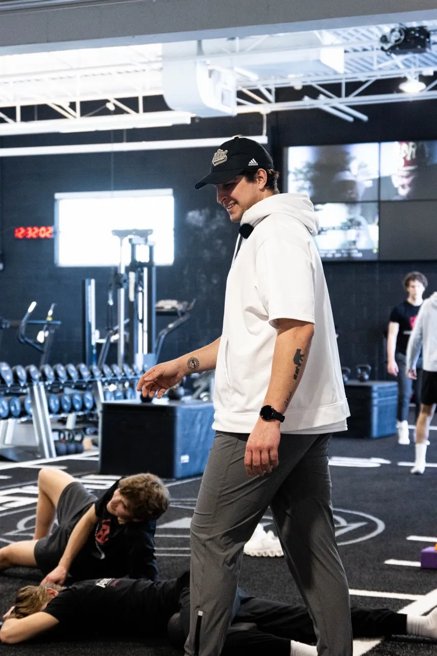 Man in white hoodie and cap walking in gym with two people lying on the floor and others in background.