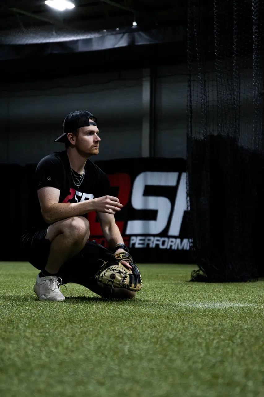 Man wearing a black cap and glove squatting on artificial turf inside an indoor sports facility.