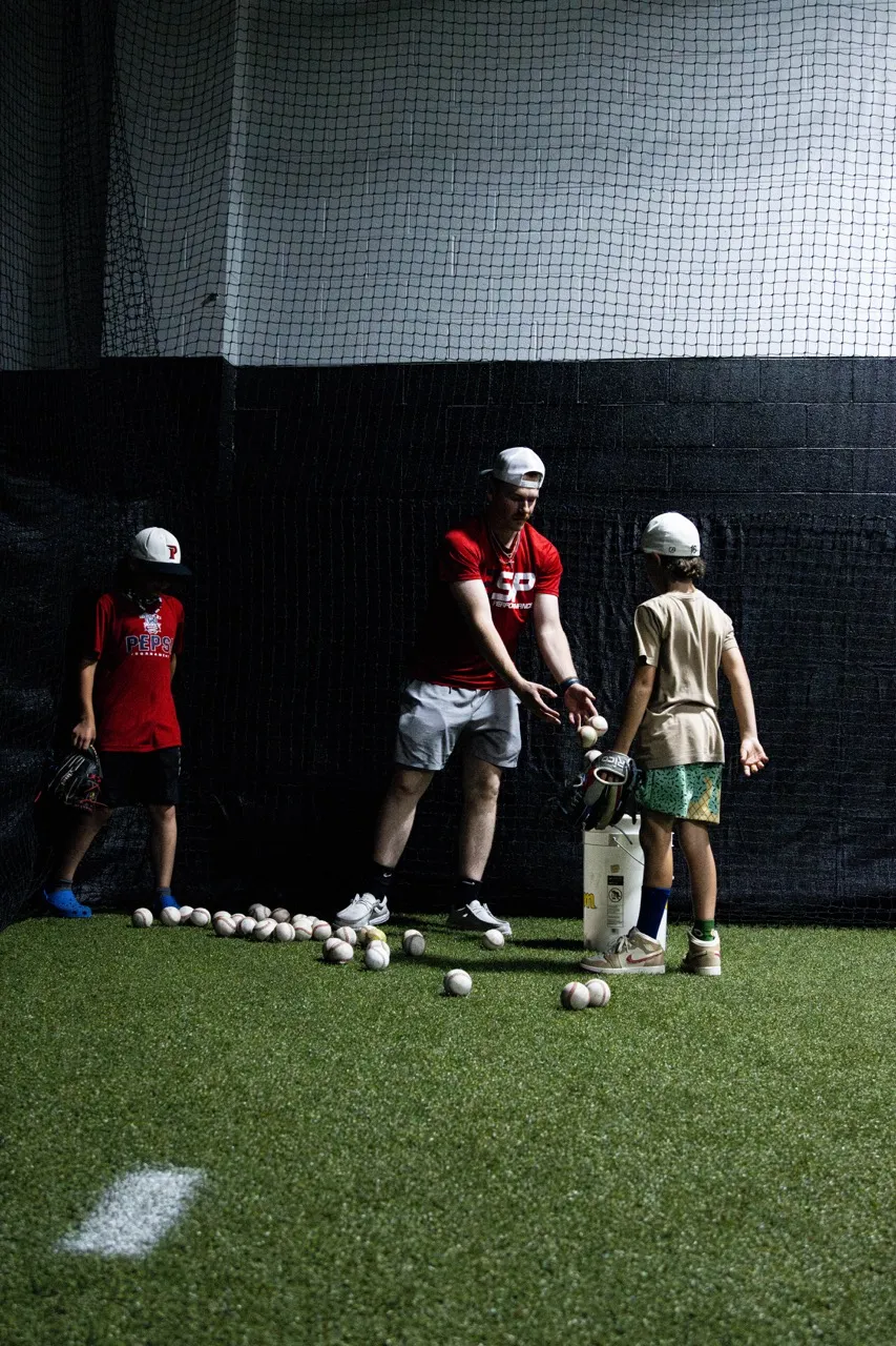 A youth baseball coach running a soft-toss or underhand fielding drill inside an indoor training facility, likely an academy like Elite Baseball Training.