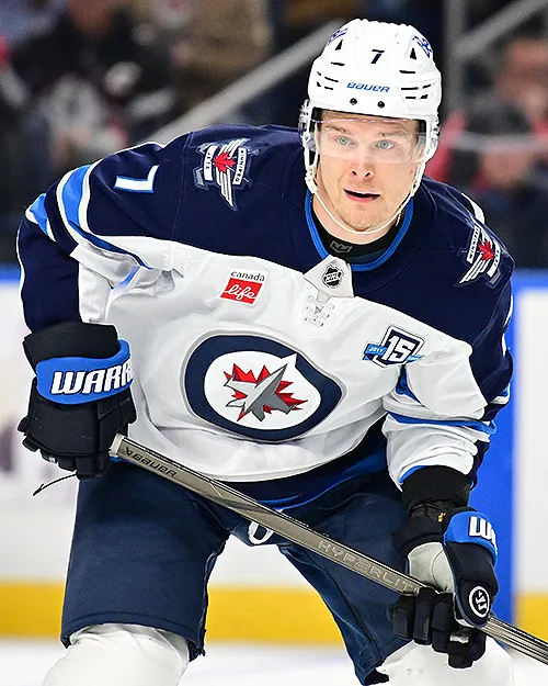 Hockey player in white Winnipeg Jets jersey with number 7 holding a hockey stick on ice.