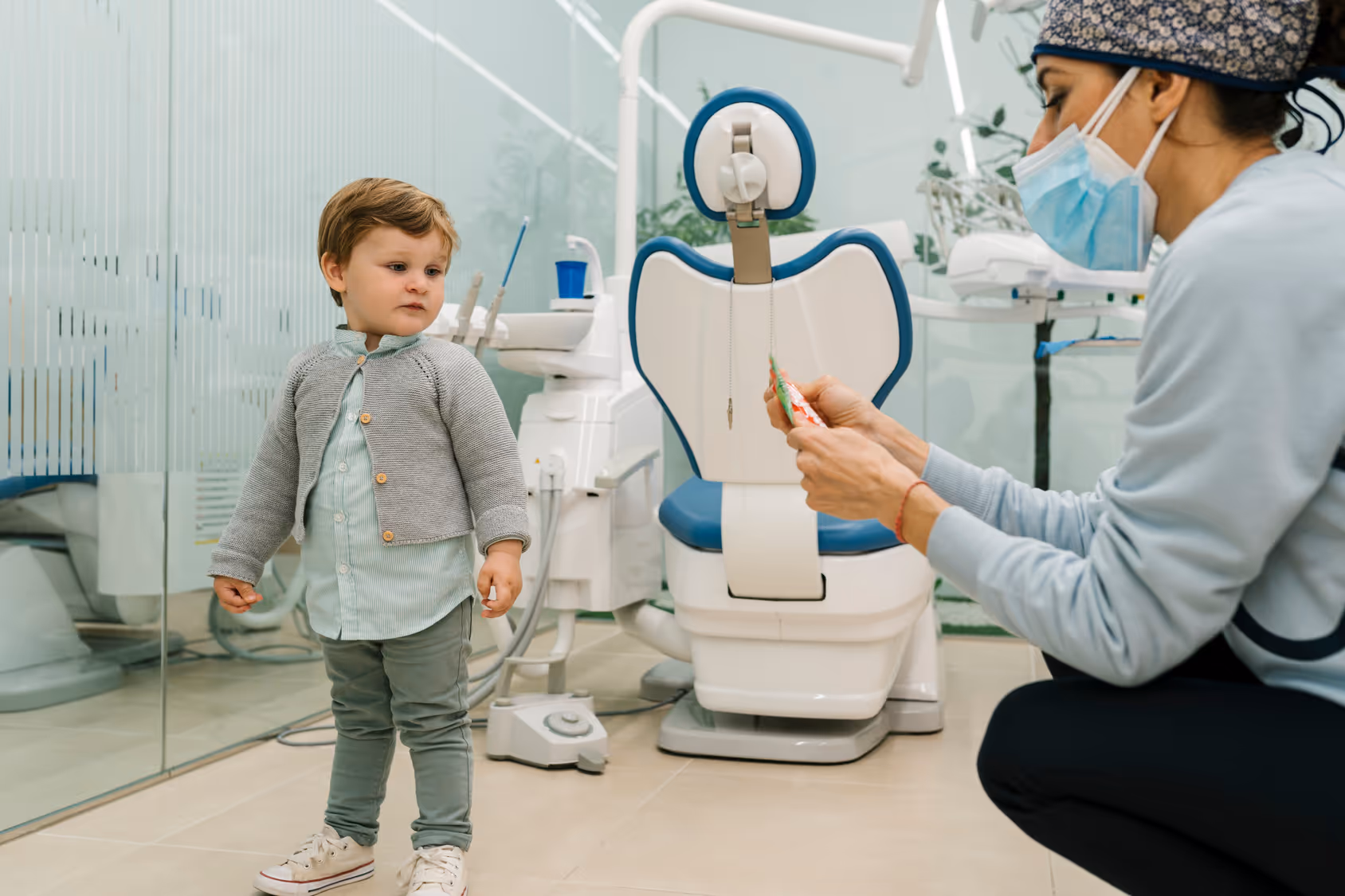 A dentist wearing a surgical mask and cap shows a tube of toothpaste to a toddler standing near a dental chair in a clinic.