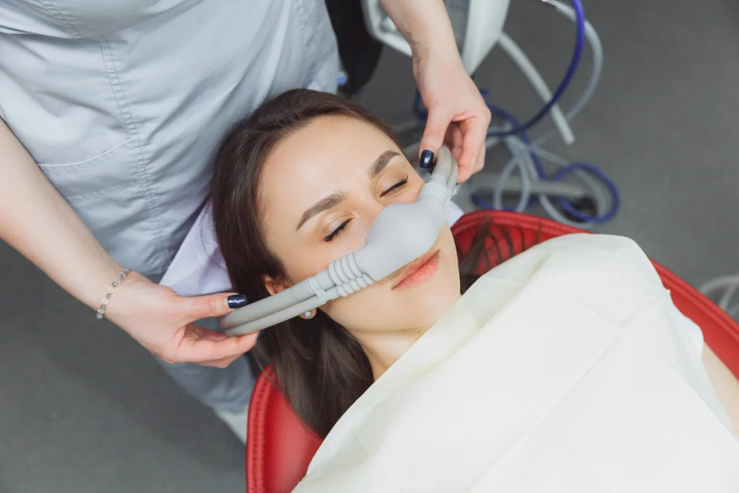 Young woman reclining with eyes closed while a healthcare professional adjusts a nasal mask for sedation or oxygen.