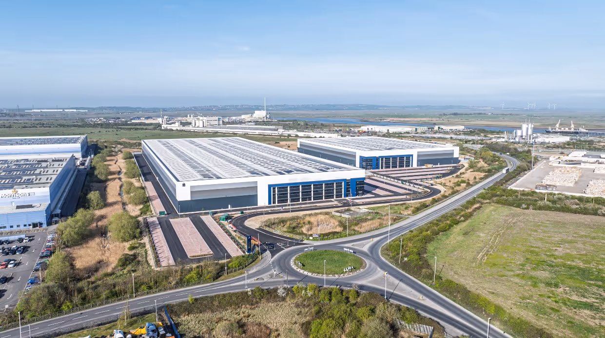 Aerial view of large industrial warehouses with solar panels on the roofs, surrounded by roads, greenery, and distant industrial facilities under a clear blue sky.