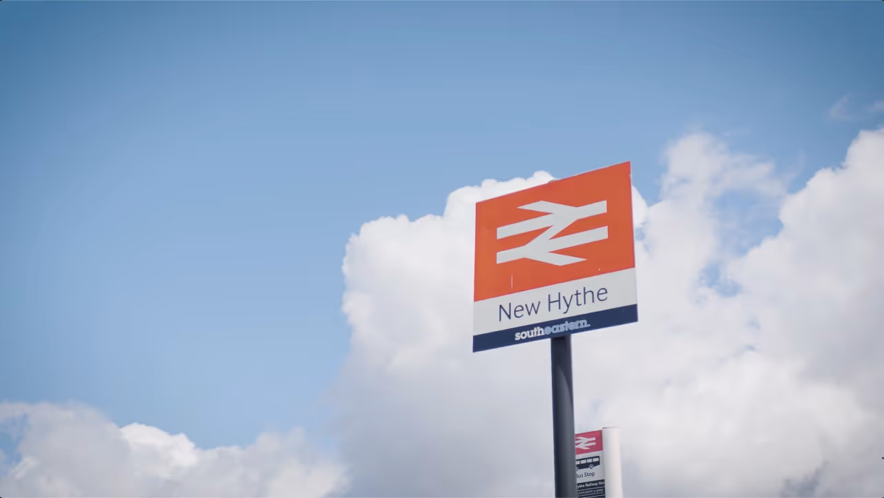 New Hythe train station sign under a blue sky with clouds.