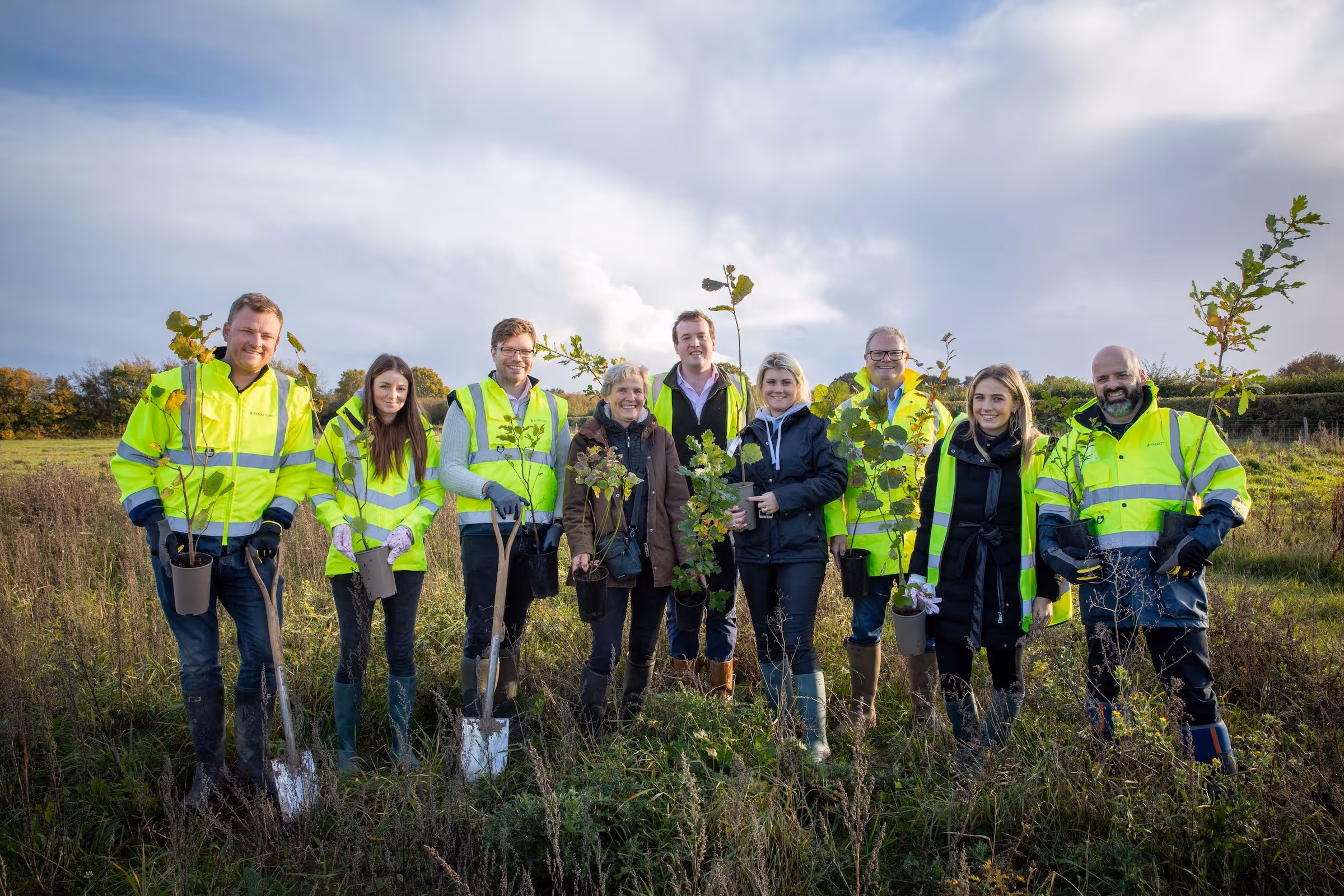 Group of nine people outdoors in reflective jackets and boots holding small trees and shovels ready for planting.