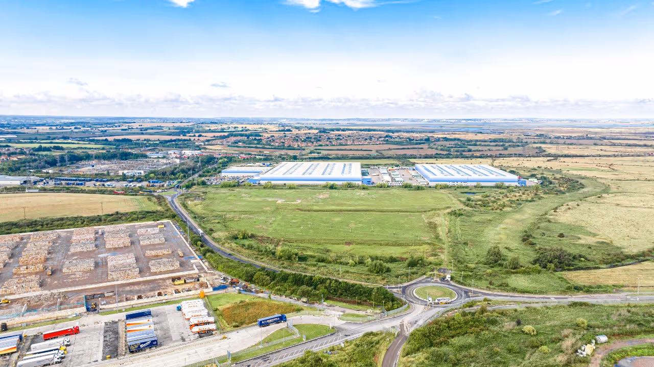 Aerial view of an industrial area with large warehouses, trucks, and a roundabout surrounded by green fields under a blue sky.